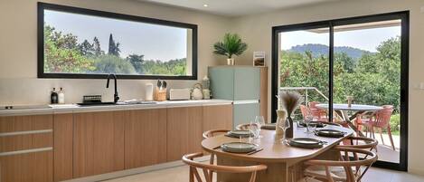 A modern kitchen with a dining area, large windows, and a view of greenery.