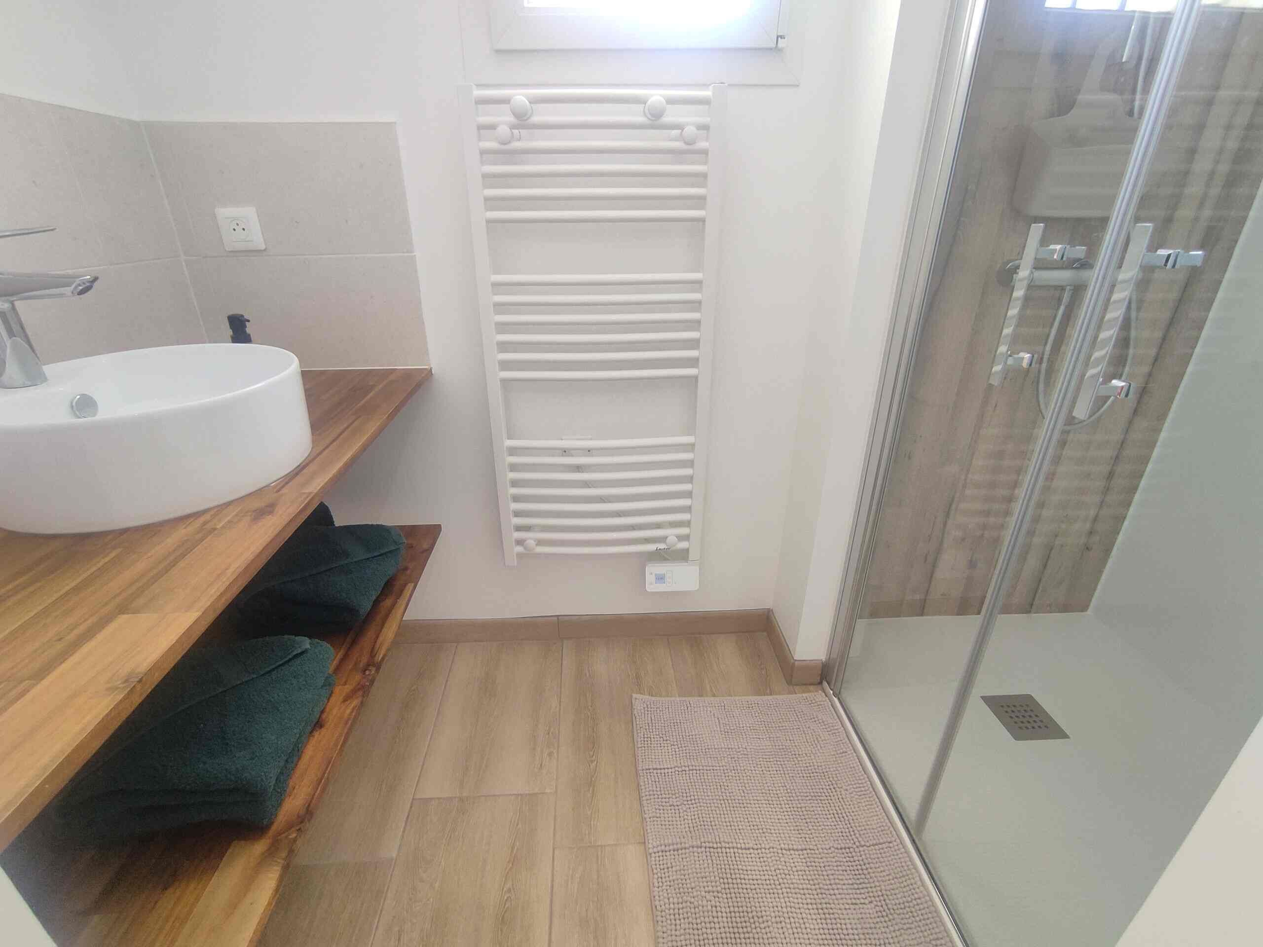 Een badkamer met een witte wastafel, houten planken, een handdoekhouder, een douchecabine van glas en een houten vloer.