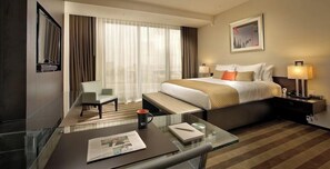 Ein Hotelzimmer mit einem großen Bett, einem Schreibtisch mit Stuhl, einem Glastisch, einem Nachttisch mit Lampe und einem Fenster mit Vorhängen.