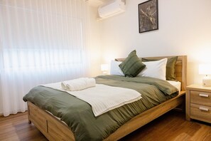 Ein ordentlich bezogenes Bett mit einem grünen Überwurf, weißen Kissen und einem weißen Handtuch. Ein Nachttisch mit Lampe und Schublade steht daneben. Der Raum hat Holzboden und ein Fenster mit durchsichtigen Vorhängen.