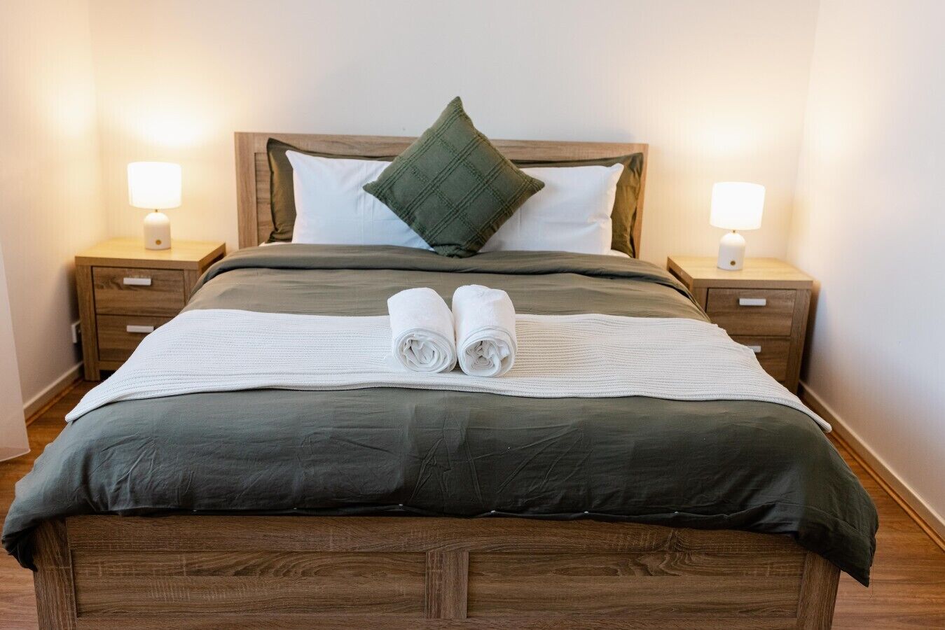 A neatly made bed with a dark green bedspread, two white pillows, a folded white towel, and a green throw pillow. There are two bedside lamps on wooden nightstands.