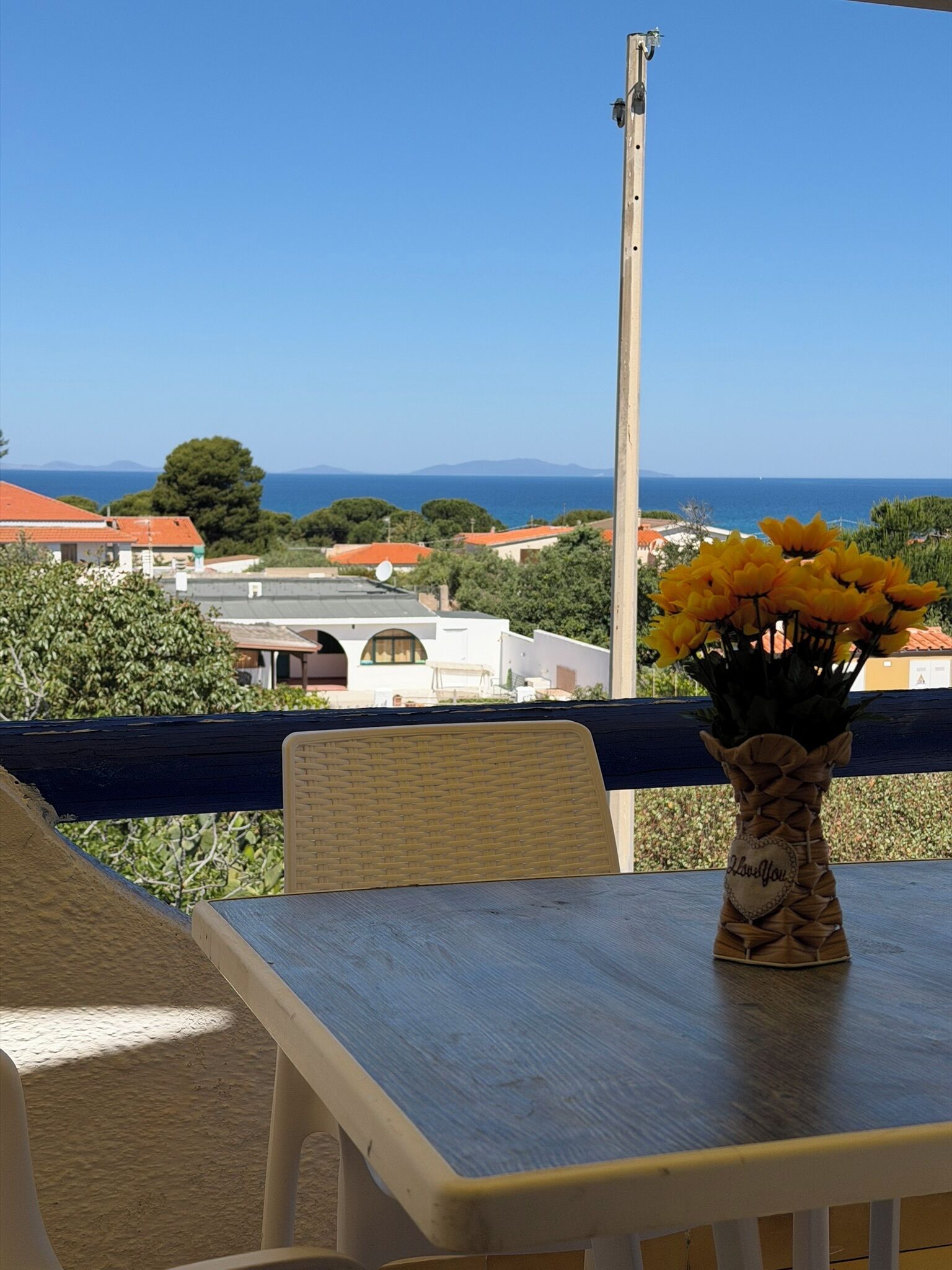A balcony with a wicker chair and a table, a vase with yellow flowers, and a view of houses and the sea.