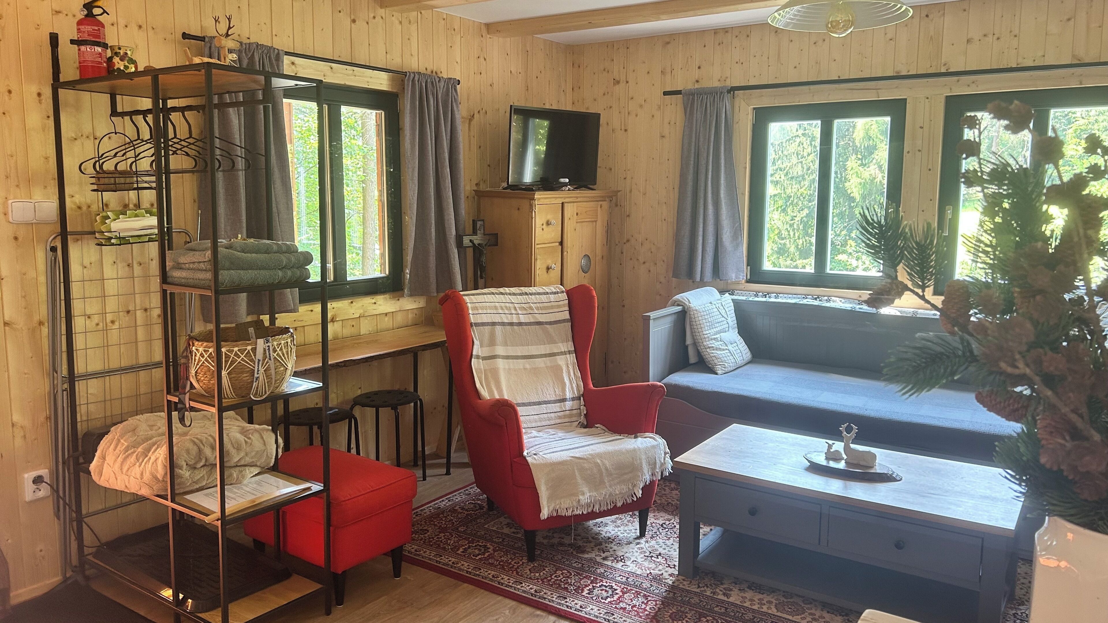 A cozy cabin interior with a red armchair, a blue sofa, a wooden TV stand, and a shelf with towels and baskets.