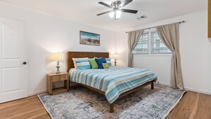 A bedroom with a bed, two bedside lamps, a ceiling fan, a window with curtains, and a painting on the wall. - Game Room | Open Living Space | Large Kitchen | Sunroom | Fenced Backyard | Fire Pit (St. Simons Island)
