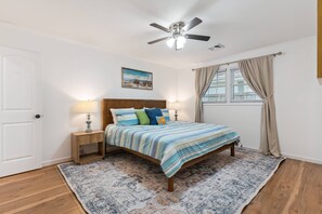 A bedroom with a bed, two bedside lamps, a ceiling fan, a window with curtains, and a painting on the wall. - Game Room | Open Living Space | Large Kitchen | Sunroom | Fenced Backyard | Fire Pit (St. Simons Island)