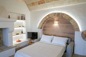 A bedroom with a stone arch, a television, a bed with white bedding, and shelves with books and decorative items. - A little gem: ancient country house with pool (Leuca)