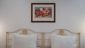 Una cama doble con ropa blanca y un cabecero de madera, un cuadro floral enmarco en la pared superior, y una pared lisa.