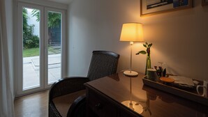 Una habitación con un escritorio de madera, una silla, una lámpara, un cuadro en la pared y vistas a través de una puerta de cristal.