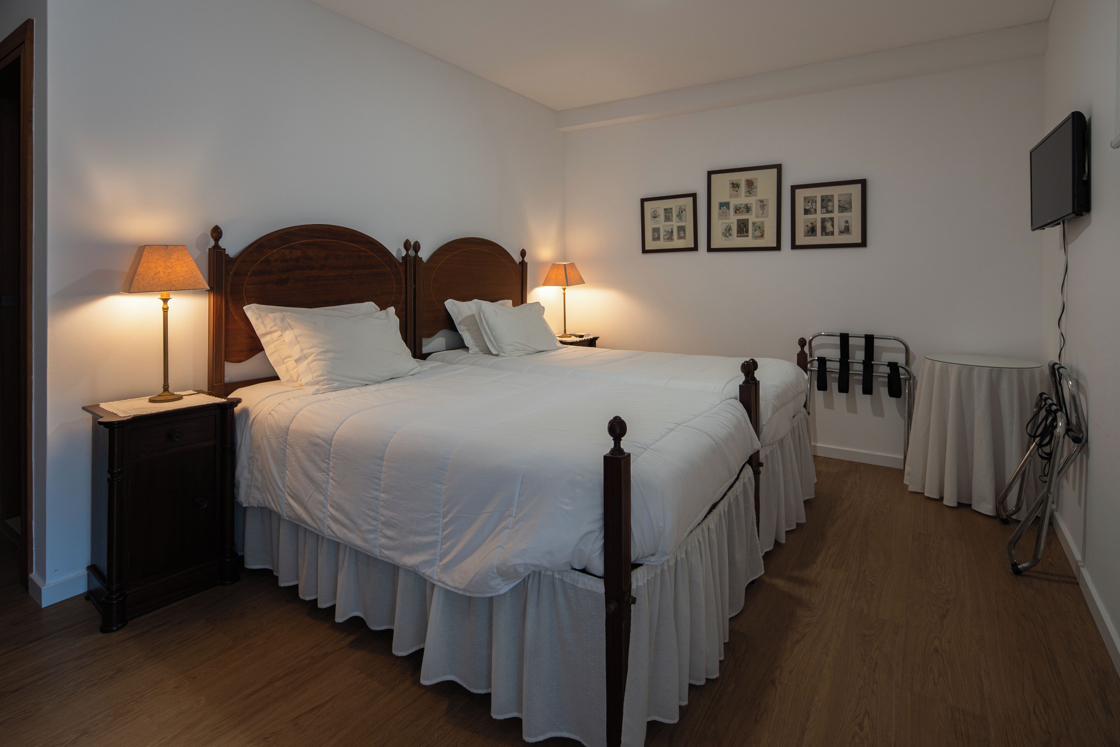 A neatly made bed with a wooden headboard, two bedside tables with lamps, a wall-mounted TV, and a set of framed pictures.