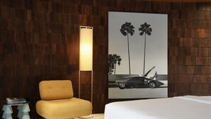 A hotel room with a bed, a chair, a lamp, a painting of a car and palm trees, and a small table with books. - Nya Uluwatu (Pecatu)