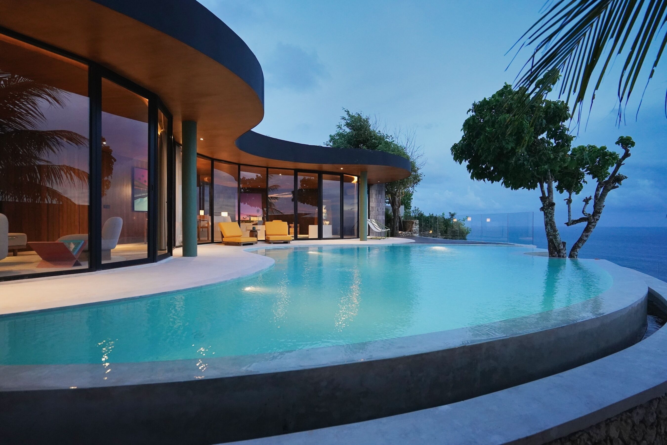 Una piscina infinita moderna con borde curvo, rodeada de mobiliario exterior y un área con paredes de vidrio.
