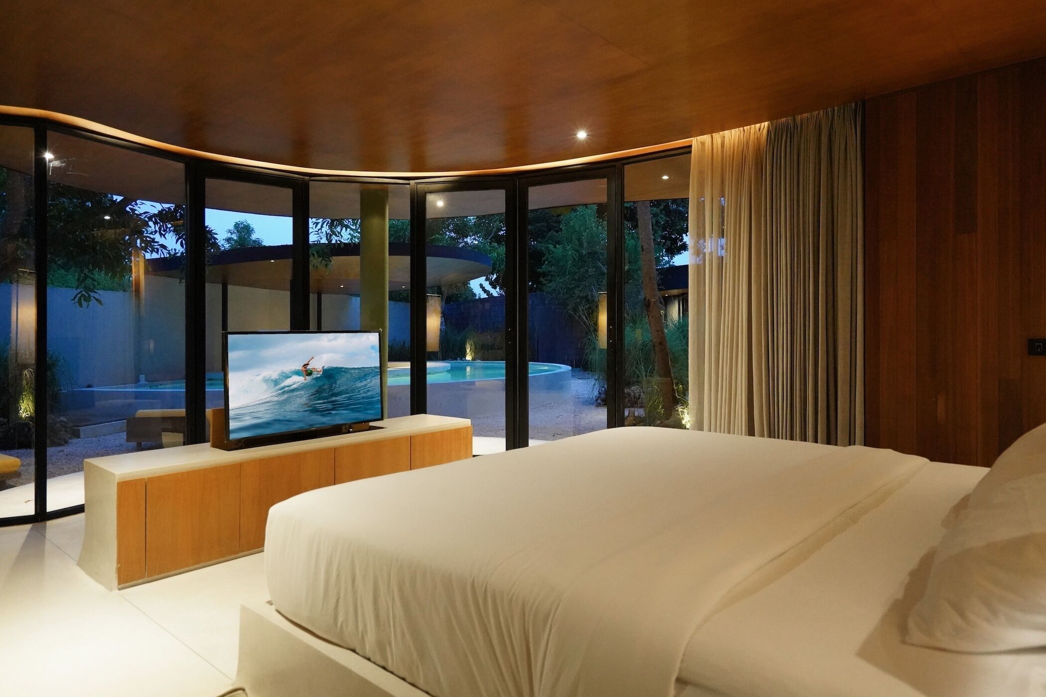 A modern bedroom with a large bed, a television on a wooden console, and a view of an outdoor swimming pool.