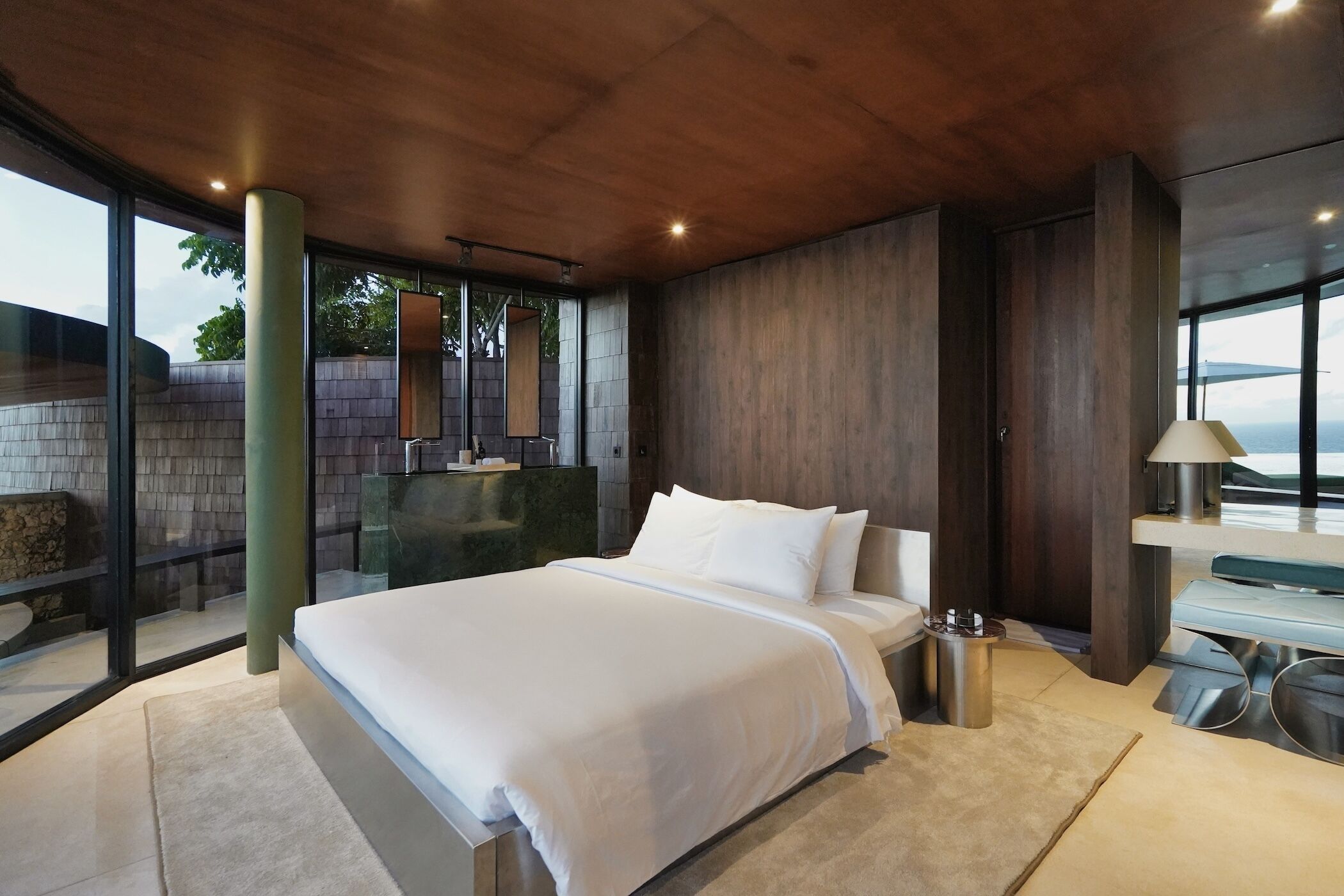 A modern bedroom with a large bed, a glass wall, and a view of the outdoors.