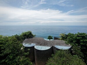 A modern building with a unique curved roof design, surrounded by lush greenery and overlooking a calm sea. - Nya Uluwatu (Pecatu)