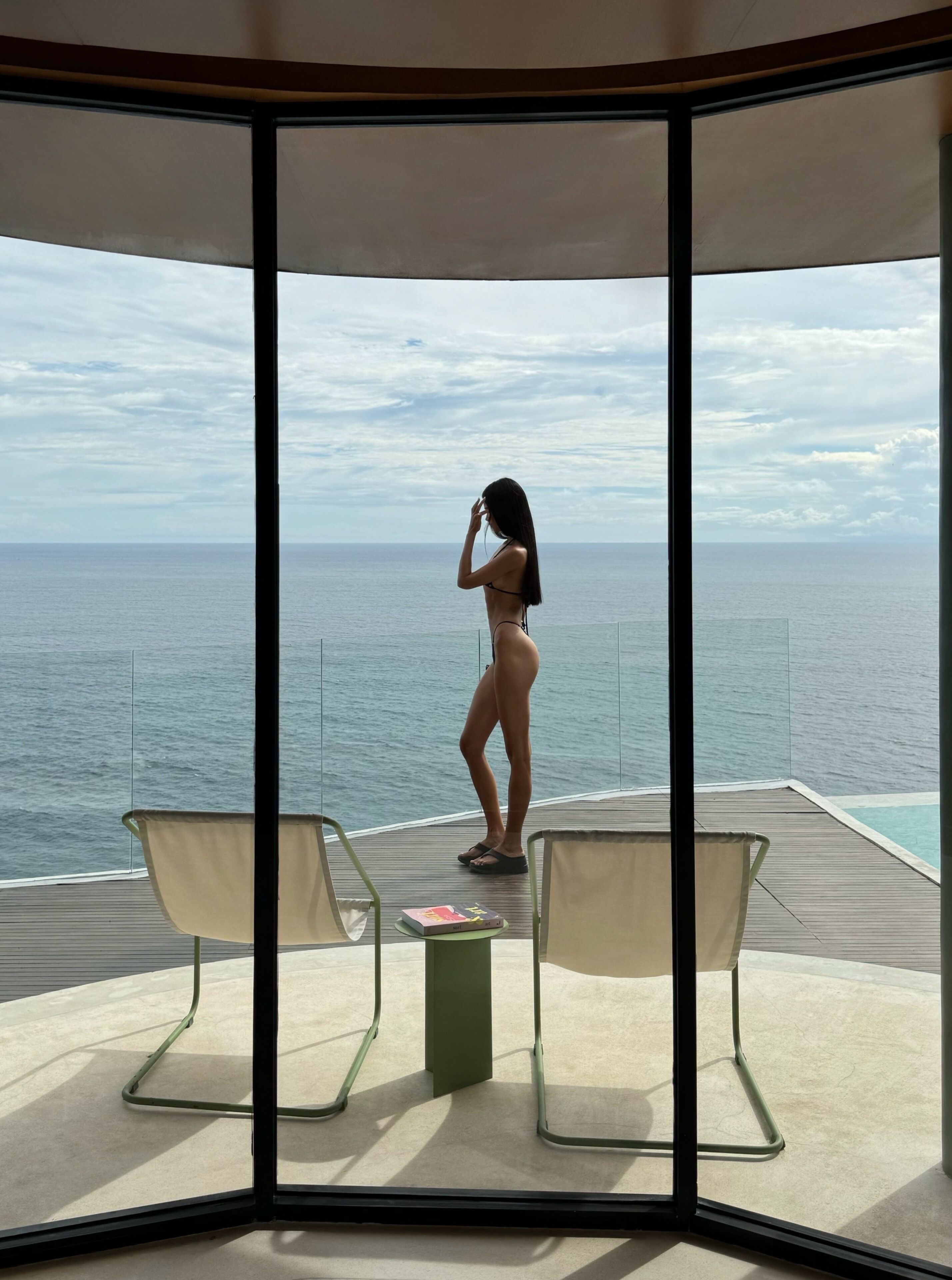 A room with a large window overlooking the sea, two chairs, and a small table with a book.