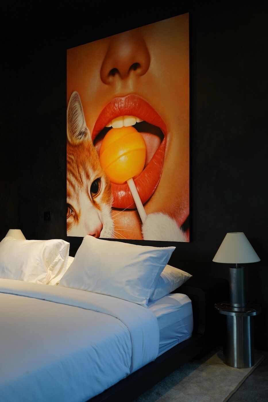 A bed with white linens, a large painting on the wall, and a lamp on a side table.