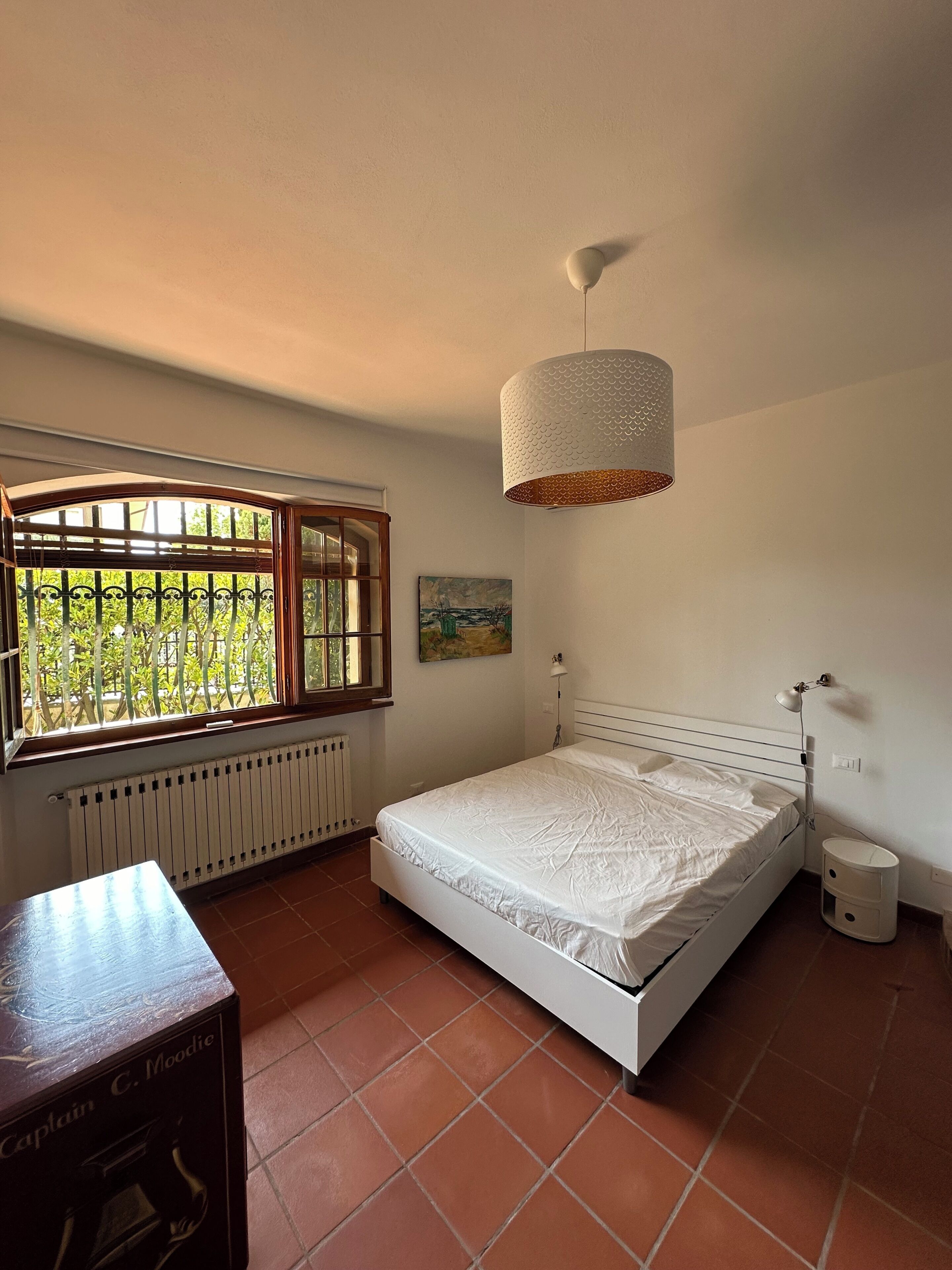 Une chambre à coucher avec un lit, une fenêtre, un radiateur, une lampe et une petite table.