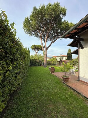 Un jardín bien cuidado con un árbol grande, un seto y una zona de patio con mobiliario exterior.