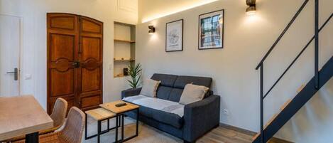 A living room with a sofa, a dining table, chairs, a staircase, and a wall-mounted air conditioner.