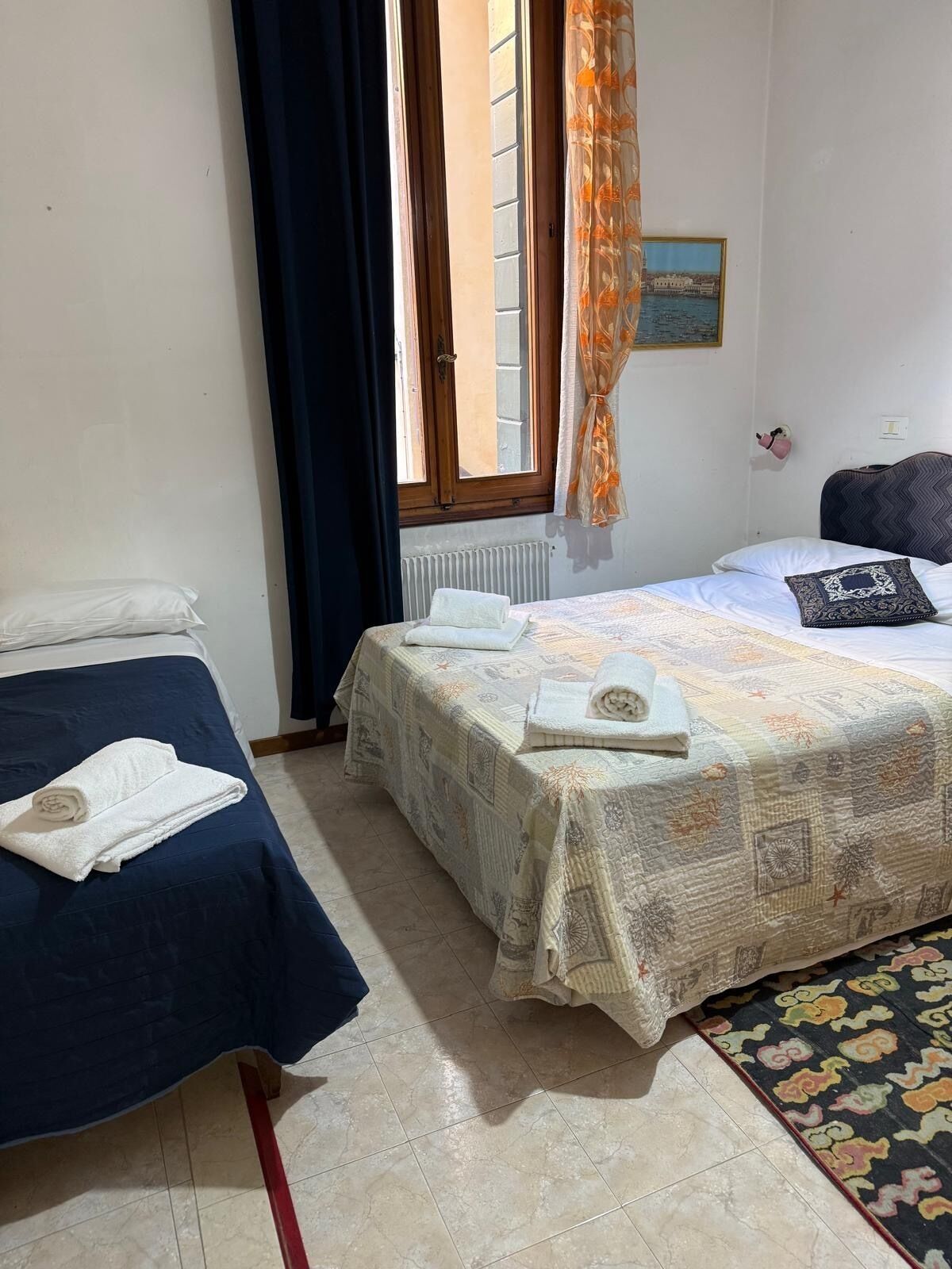 Une chambre avec deux lits, une fenêtre avec des rideaux, un radiateur et un petit tableau accroché au mur.