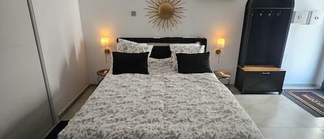A bedroom with a bed, two bedside lamps, a wall-mounted sun decoration, an air conditioning unit, and a door with a handle.