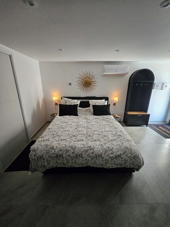A bedroom with a bed, two bedside lamps, a wall-mounted sun decoration, an air conditioning unit, and a door with a handle.