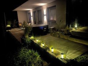 Una casa moderna con un área de estar cubierta al aire libre, un jardín pequeño con plantas y un sendero que conduce a la entrada.
