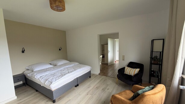 A bedroom with a bed, a chair, a bedside table, a wall-mounted lamp, and a door leading to another room.
