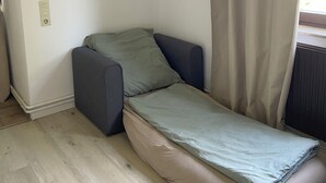 A small, single-bed room with a wooden floor, a grey sofa, a beige mattress, and a window with curtains.