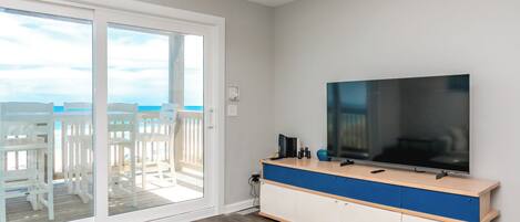 A room with a large flat-screen TV on a wooden and blue cabinet, a sliding glass door leading to a balcony with white chairs, and a small table with a decorative item.