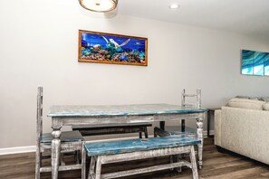 Un coin repas avec une table et des bancs en bois rustique, un tableau encadré au-dessus, et un canapé à l’arrière-plan.