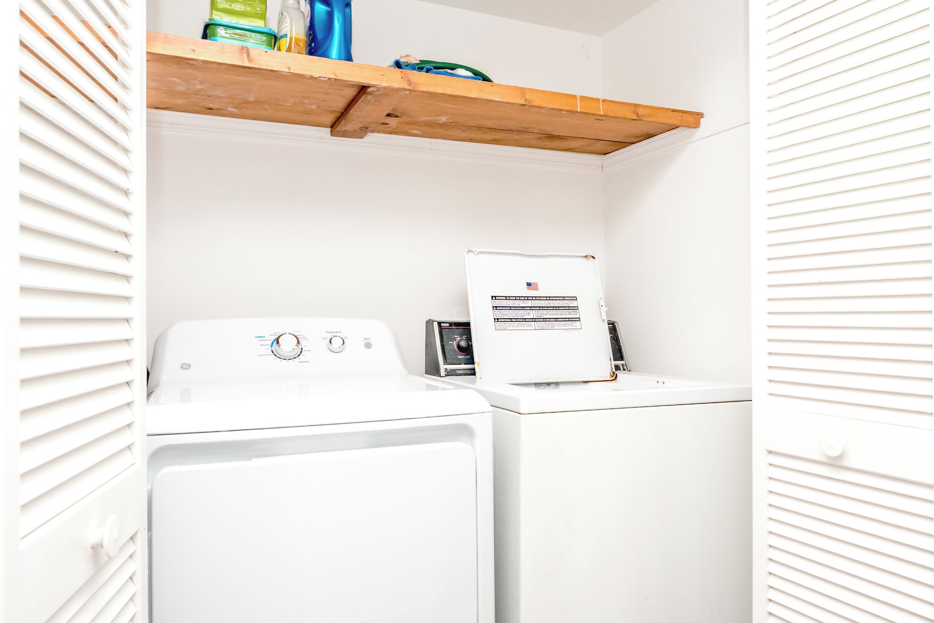 A laundry room with two white washing machines and a wooden shelf above them.