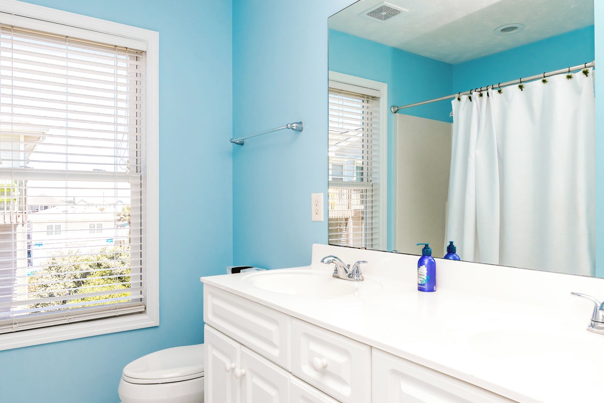 Une salle de bain avec un meuble-lavabo blanc, un miroir, des toilettes et une fenêtre avec des stores.