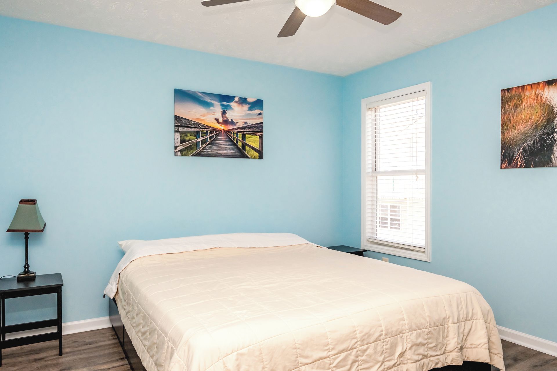 Un dormitorio con cama, mesita de noche, ventilador de techo y dos cuadros colgados en la pared.