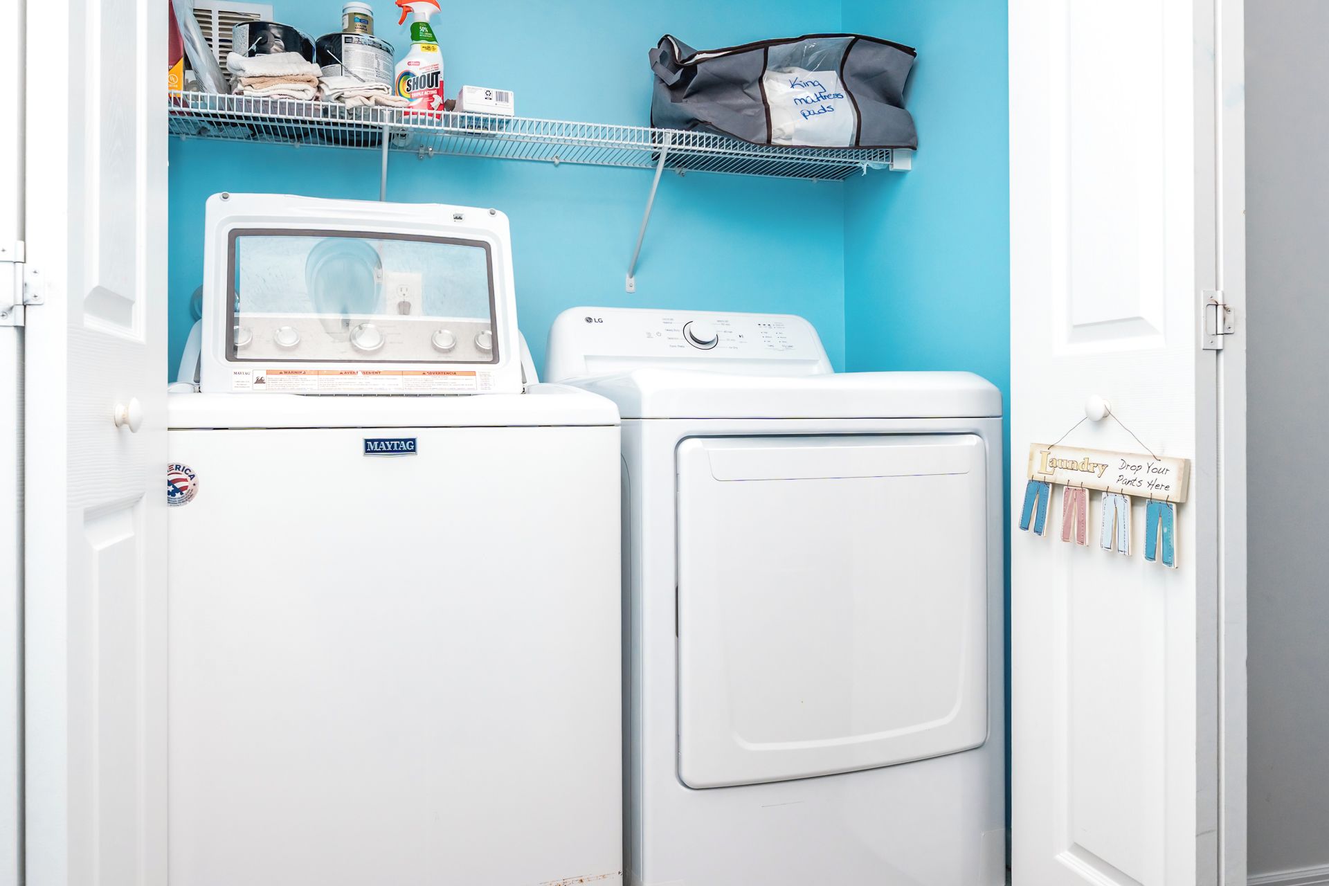 A laundry area with a Maytag washer and dryer, a shelf with cleaning supplies, and a sign indicating where to drop off laundry.