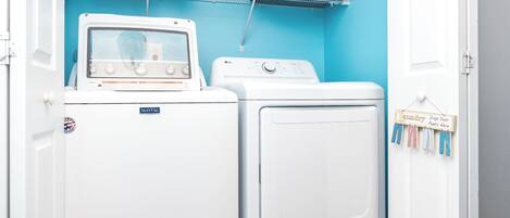 A laundry area with a Maytag washer and dryer, a shelf with cleaning supplies, and a sign indicating where to drop off laundry.