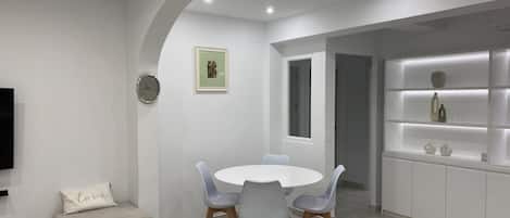 Et moderne spiseområde med et rundt bord, grå stole, en sofa og et indbygget reolsystem med dekorative genstande.