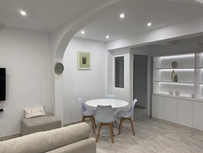 A modern dining area with a round table, grey chairs, a sofa, and a built-in shelving unit with decorative items. - Sea View Retreat - Beachfront Two-Bedroom Penthouse (Calpe)