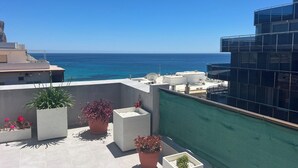 A rooftop terrace with potted plants, a view of the sea, and a modern building with reflective glass windows. - Sea View Retreat - Beachfront Two-Bedroom Penthouse (Calpe)