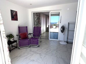 Quarto com porta de vidro deslizante para uma varanda, um ventilador e uma cadeira roxa com almofada vermelha.