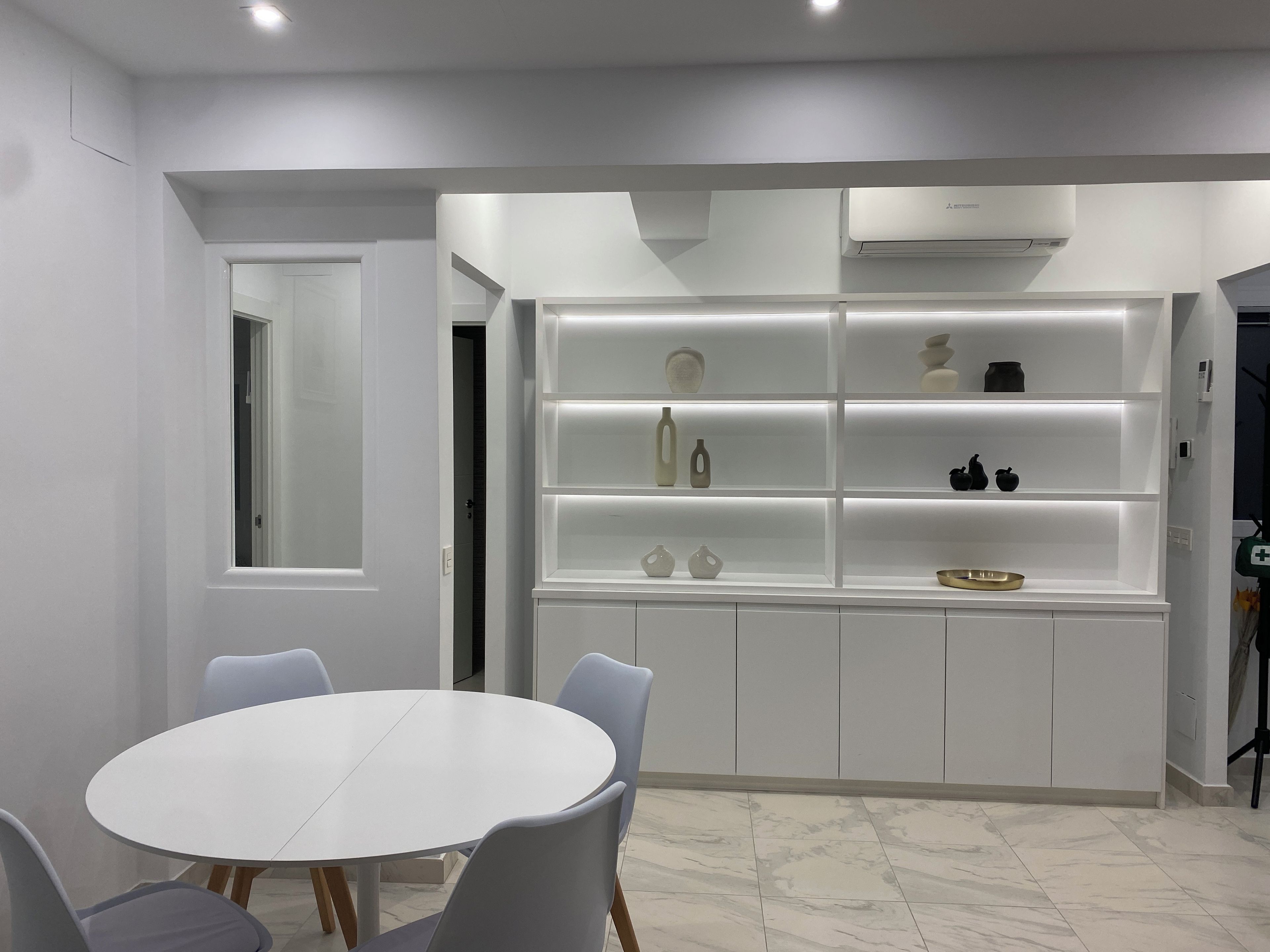 A modern kitchen with white cabinetry, a built-in shelf unit displaying various kitchenware, and a round dining table with chairs.