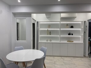 Uma cozinha moderna com armários brancos, uma estante embutida exibindo utensílios de cozinha e uma mesa de jantar redonda com cadeiras.