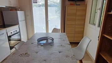 Une cuisine compacte comprenant une table à manger, des chaises, un réfrigérateur, un téléviseur et une porte-fenêtre coulissante donnant sur l’extérieur.
