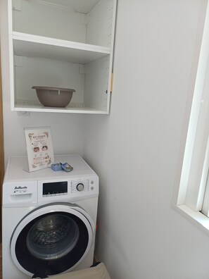 Uma área de lavanderia compacta, com máquina de lavar, prateleira acima e janela à direita.
