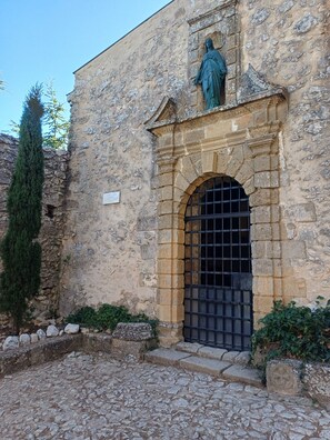 Un edificio in pietra con una statua sopra un portale, un sentiero in pietra e un alto abete sempreverde.