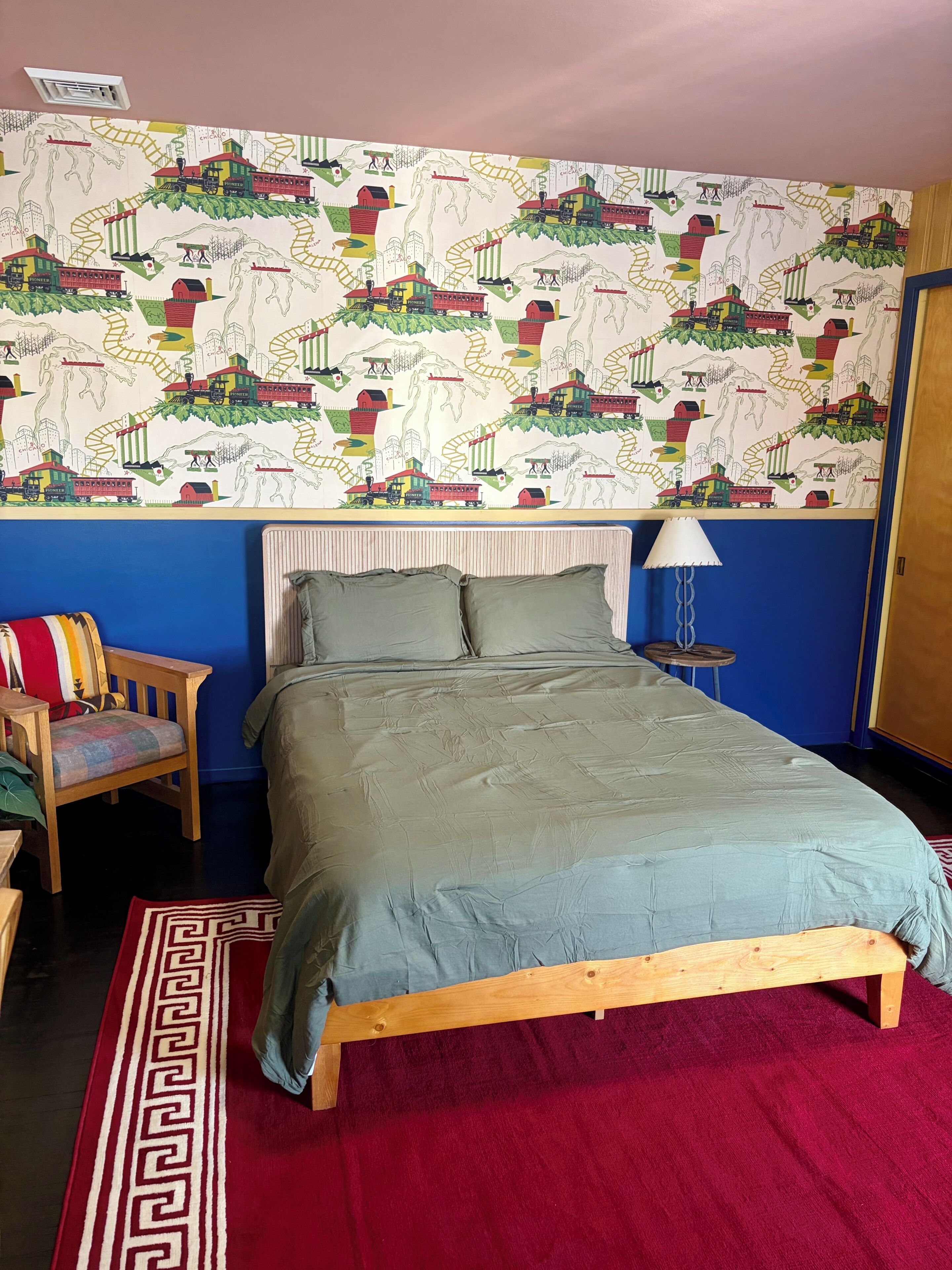 A bedroom with a bed, pillows, a chair, a lamp, and a patterned wallpaper.