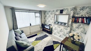 Una sala de estar con un sofá, un televisor, una estantería, un ventanal con cortinas y una pared con diseño.