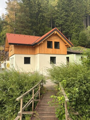 A wooden cabin with a red roof, a wooden deck, and a forested backdrop.