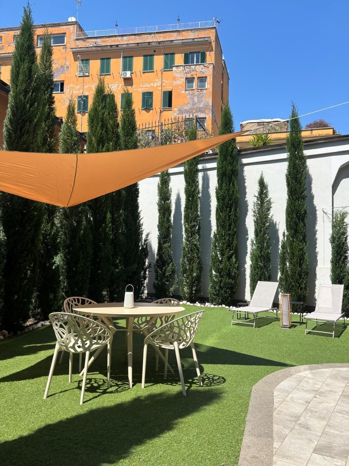 An outdoor patio with a round table and chairs, surrounded by artificial grass and tall evergreen trees.