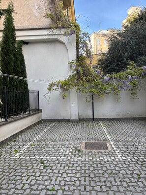 A courtyard with a cobblestone floor, a black metal fence, and a building with a white wall and purple flowering vines. - Apartment with a private garden and a private parking space (Roma)